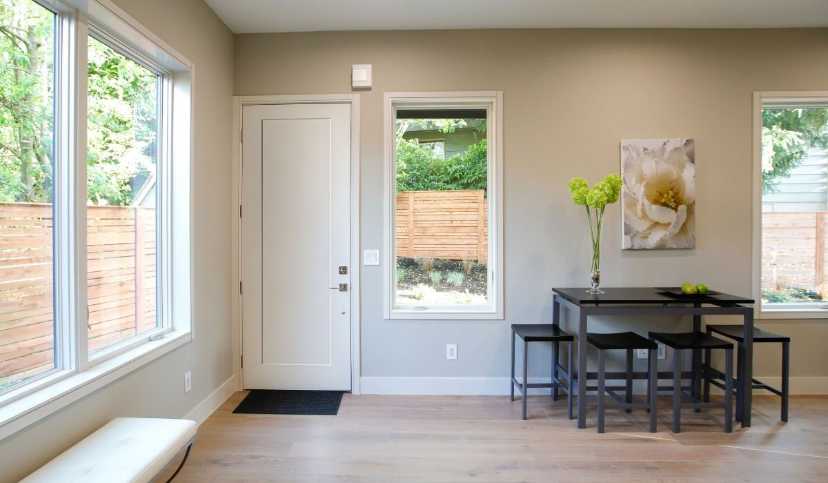 westguard-collection-homepage Bright, minimalist entryway with a white front door, black-framed windows, light hardwood floors, and a small black dining table with four stools set against a taupe wall decorated with a floral print and a vase of greenery.